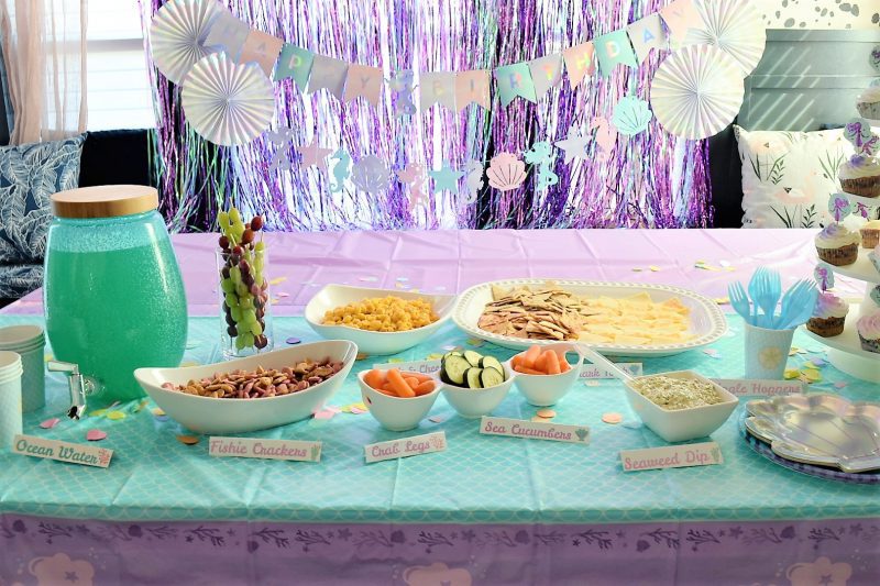 under the sea mermaid party food and party decorations with happy birthday banner and mermaid themed backdrop